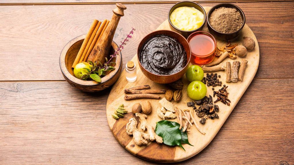 A wooden platter displays various spices, herbs, and ingredients, including cinnamon sticks, green apples, and a bowl of dark paste.